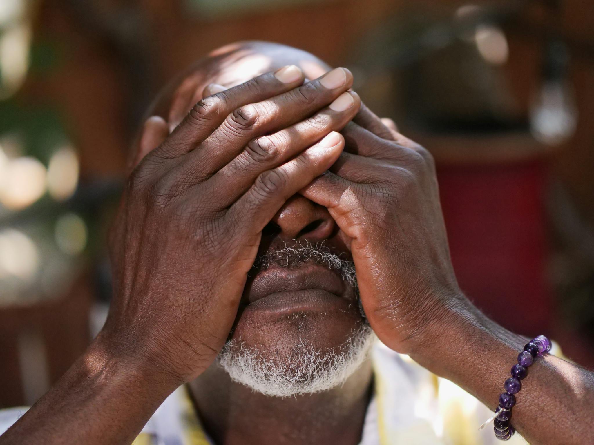 Close-up of an elderly man with a beard covering his face with his hands outdoors.